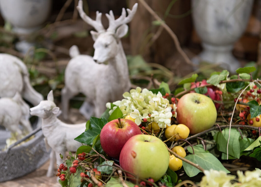 Eine Nahaufnahme eines farbenfrohen Herbstgestecks mit roten und grünen Äpfeln, gelben Beeren und weißen Blumen, mit zwei weißen Hirschstatuen im Hintergrund. Üppige grüne Blätter umgeben das Obstarrangement.