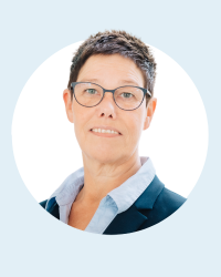 Eine Person mit kurzen braunen Haaren und Brille, die einen blauen Blazer und ein hellblaues Hemd mit Kragen trägt, vor hellblauem Hintergrund.
