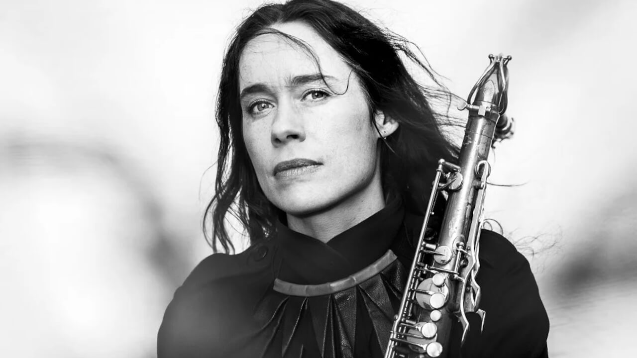 A black and white photo of a woman with long hair, wearing a dark top, holding a saxophone and looking thoughtfully at the camera. The background is soft and blurred.