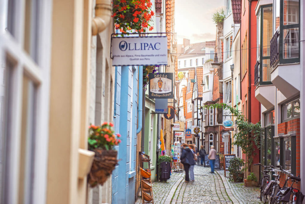 Eine schmale Straße im Bremer Schnoorviertel mit Kopfsteinpflaster, gesäumt von bunten Gebäuden, Ladenschildern und Blumenkörben. Ein paar Leute gehen die Straße entlang, und die Atmosphäre ist lebendig und charmant, wie in einer malerischen europäischen Stadt.