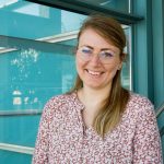 Eine Frau mit langen blonden Haaren, die eine Brille und eine rosa geblümte Bluse trägt, steht lächelnd in einem Innenraum neben einem Fenster mit blau-grünem Metallrahmen.