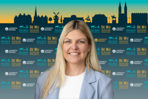 Eine lächelnde Frau mit blonden Haaren und einem hellblauen Blazer steht vor einer Kulisse mit Silhouetten der Stadt Skyline und Logos des DeWu-Kongresses in Bremen.