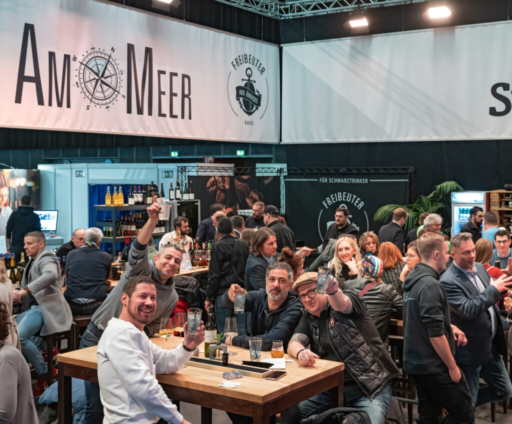 Besuchende auf der Gastro Ivent, die an einem Stand sitzen und der Kamera zuprosten.