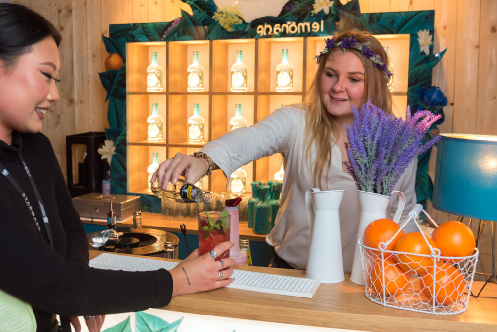 Ausstellerin mit Blumenschmuck am Kopf schenkt Besucherin am Stand einen Cocktail ein