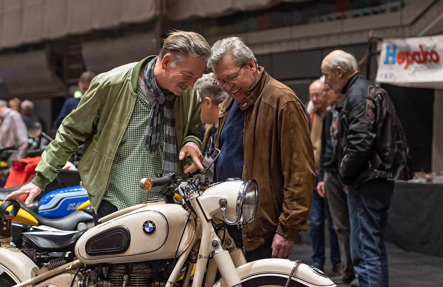 Zwei Männer betrachten lachend ein cremefarbenes, klassisches BMW-Motorrad auf der Bremen Classic Motorshow. Im Hintergrund stehen weitere bunte Oldtimer-Motorräder und interessierte Besucher in einer Messehalle. Die Stimmung ist lebendig und von Begeisterung für historische Fahrzeuge geprägt.