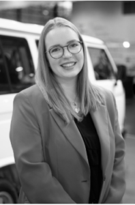 Eine Frau mit langem, glattem Haar und Brille lächelt in die Kamera. Sie trägt einen hellen Blazer über einem dunklen Oberteil und steht vor einem weißen Lieferwagen in einem Innenbereich. Das Bild ist schwarz-weiß.