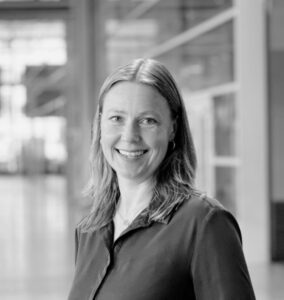 A woman with shoulder-length hair, wearing a dark blouse, smiles at the camera. The background shows a modern, glass-filled indoor space with soft natural light. The image is in black and white.