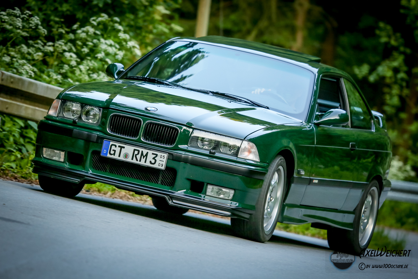Ein dunkelgrünes, sportlich wirkendes BMW E36 Coupé mit deutschem Frontkennzeichen GT-RM 3 fährt auf einer kurvigen, von Bäumen gesäumten Straße und feiert inmitten von üppigem Grün die Klassiker-Stars aus Japan.