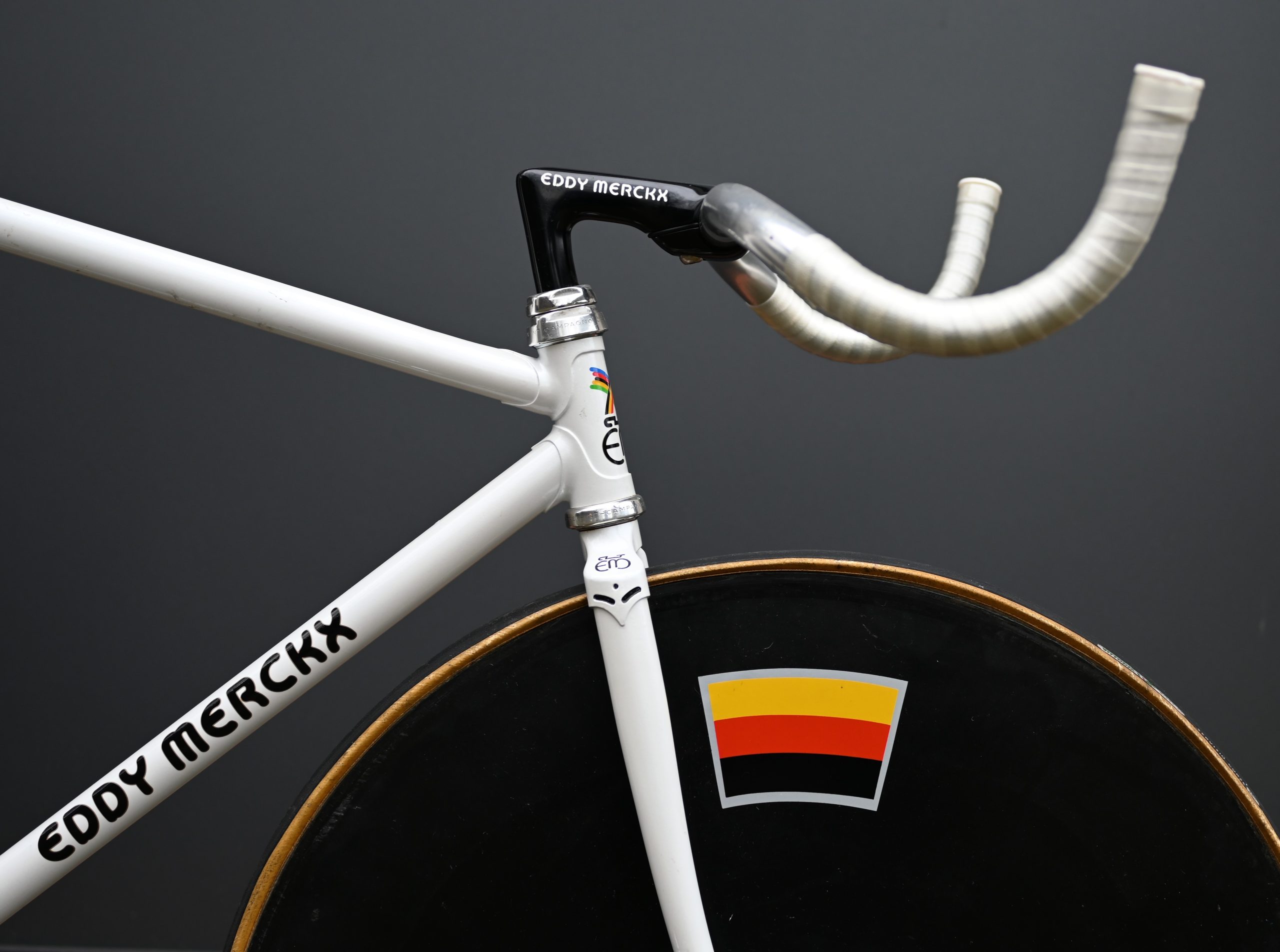 Nahaufnahme eines weißen Eddy-Merckx-Fahrrads mit markantem schwarzem Lenker und einem vorderen Scheibenrad mit einem horizontalen schwarz-rot-gelb gestreiften Aufkleber vor einem schlichten grauen Hintergrund - Bahnräder sind extreme Spezialisten.