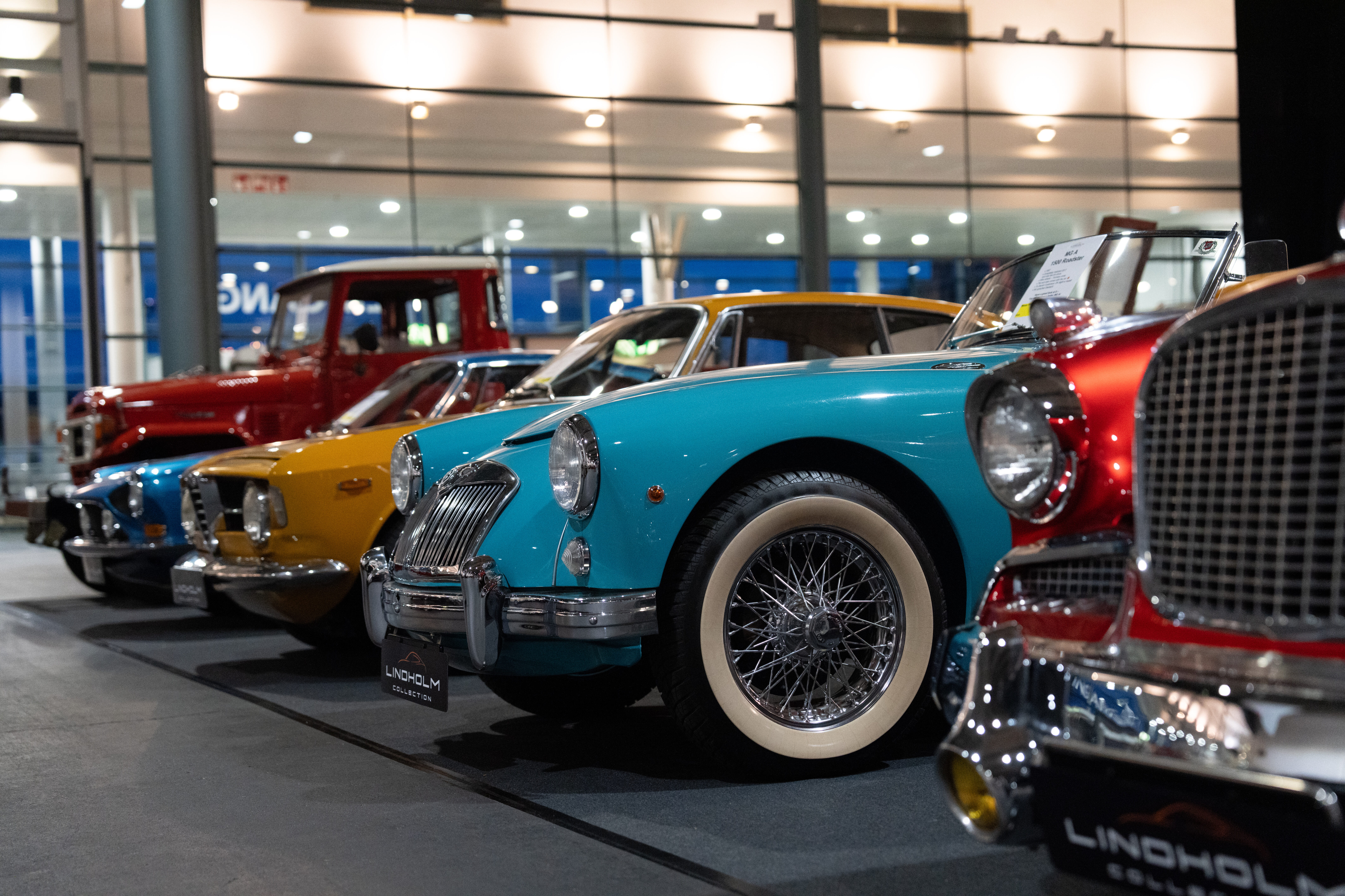 Eine Reihe von Oldtimern in verschiedenen Farben ist im Innenraum ausgestellt, mit großen Fenstern im Hintergrund und sanfter Beleuchtung, die die Details jedes Fahrzeugs hervorhebt,