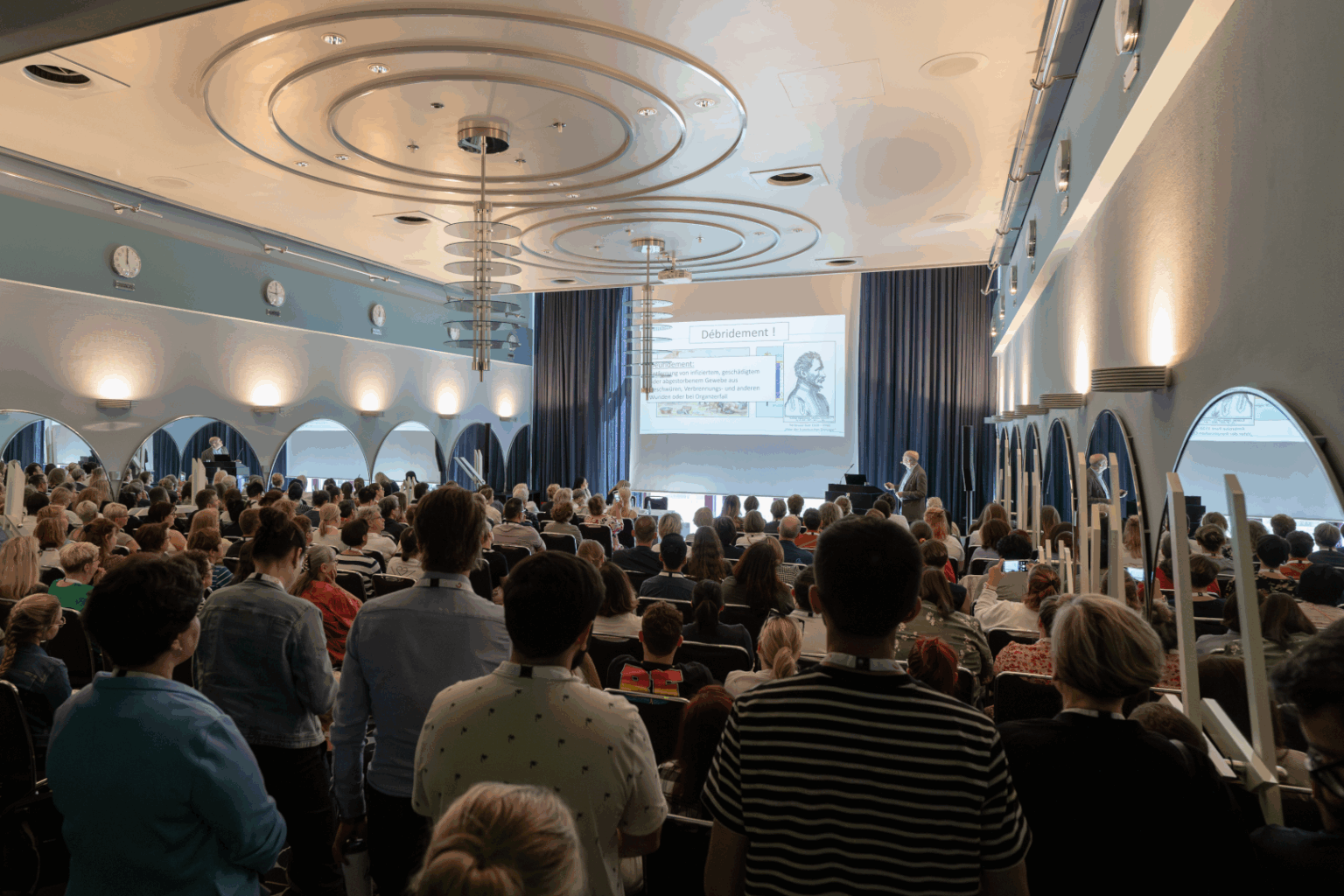 Vortragssaal beim DEWU Deutschen Wundkongress mit vielen Teilnehmenden, die einem Vortrag über Wundbehandlung folgen. Auf der Bühne spricht ein Referent vor einer Präsentation mit dem Titel „Débridement“.