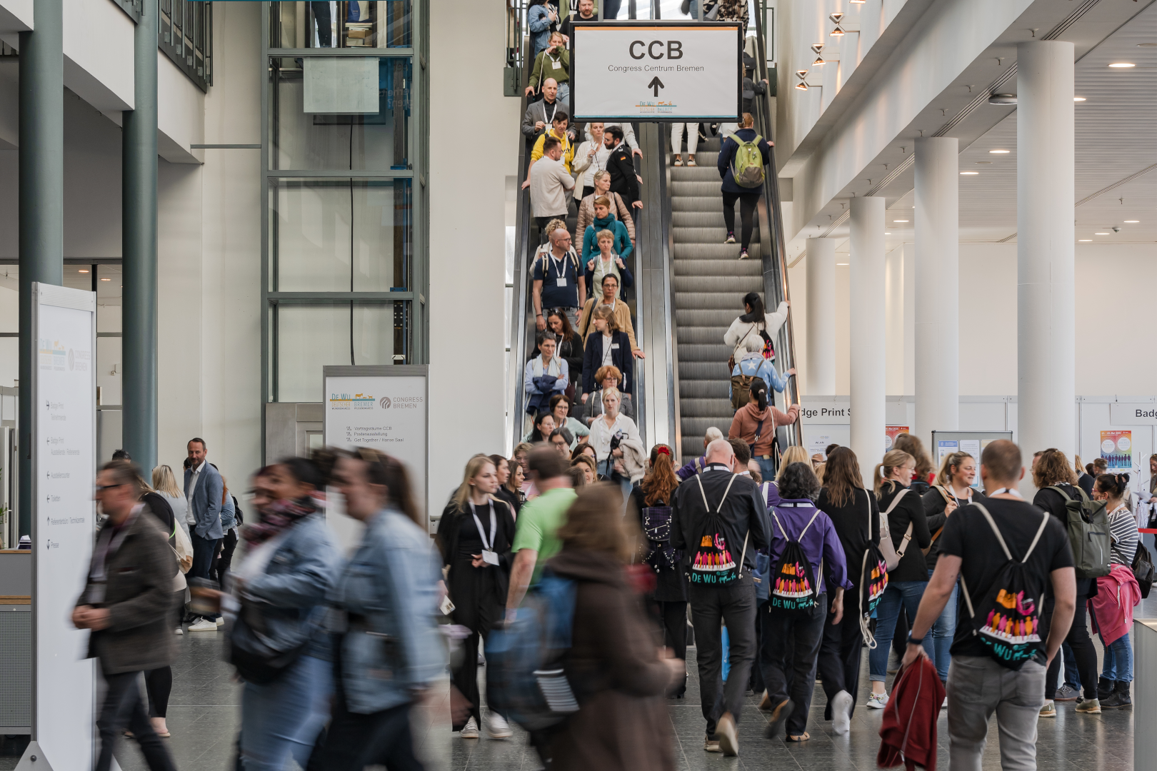 Teilnehmende des DEWU Deutschen Wundkongresses bewegen sich durch das Foyer der MESSE BREMEN, viele mit bunten DEWU-Turnbeuteln; einige fahren die Rolltreppe hinauf zu den Vortragsräumen.