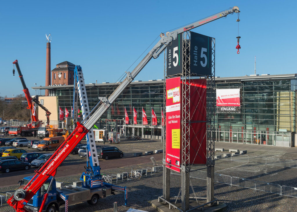 Ein großer Kran hebt ein Ausstellungsschild vor einer modernen gläsernen Messehalle mit roten Bannern an. Mehrere Kräne und Baufahrzeuge sind zu sehen, als die Menschen unter blauem Himmel in der Nähe des Eingangs der MESSE BREMEN spazieren gehen.