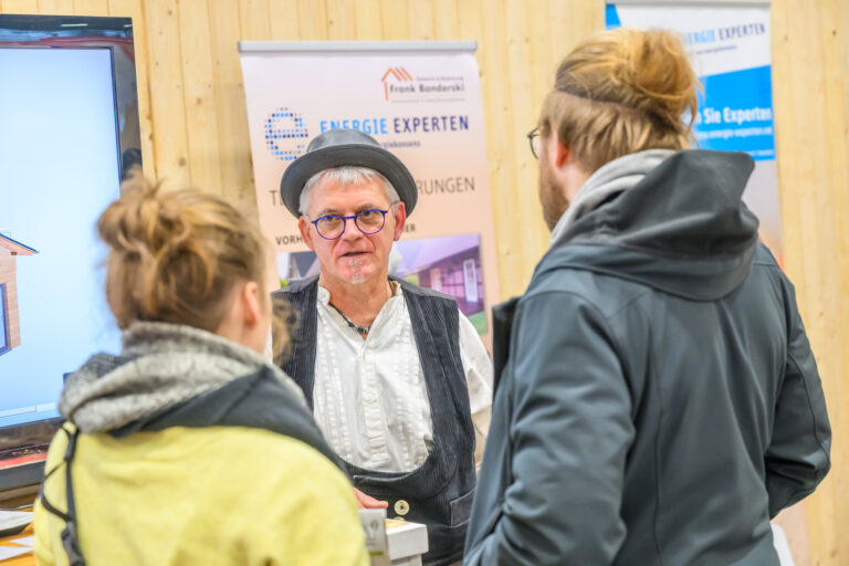 Ein Mann in traditioneller Kleidung und mit Hut spricht mit zwei Personen auf der hanseBAU & Bremer Altbautage auf der MESSE BREMEN mit Informationsplakaten im Hintergrund. Die professionelle, lehrreiche Umgebung scheint sich in einer Messehalle an einem Ausstellerstand zu befinden.